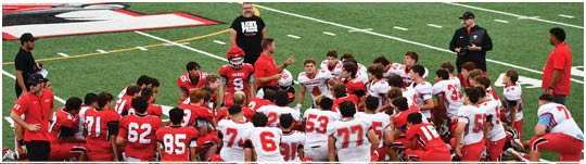 A group of people wearing red and white sports uniforms are kneeling and sitting on the sidelines of a football field. Description generated by AI