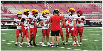 A group of people wearing red and white football uniforms and yellow helmets stand on a field while one person in a red shirt and black shorts appears to be giving instructions. Description generated by AI