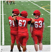 Two people wearing red padded helmets and red sports uniforms watch a group of players in red and white uniforms with yellow helmets on a football field with empty bleachers in the background. Description generated by AI