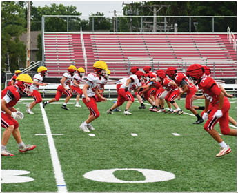 Two teams of people in red and white sports uniforms with helmets are lined up facing each other on a football field preparing for a play. Description generated by AI