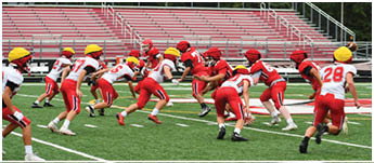 People wearing red and white sports uniforms and yellow helmets are actively playing football on a green field with white yard lines. Description generated by AI