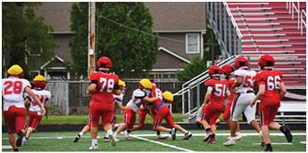 Children in red and white football uniforms and yellow helmets are playing on a grassy field near bleachers and houses. Description generated by AI