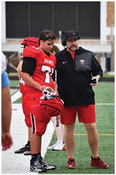 A person wearing a red football jersey with the number 71 and the word “HAWKS" stands next to another person wearing a black jacket and headset. Description generated by AI