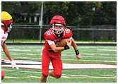 Two people in football uniforms are on a field with one person running while holding a football and the other person reaching out toward them. Description generated by AI