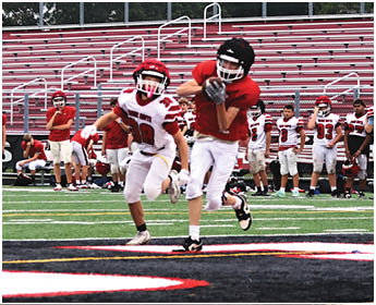 Two people in football uniforms are actively playing on a field with others standing in the background near bleachers. Description generated by AI