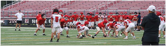 People in red and white football uniforms are lined up on a field preparing for a play while coaches and others stand on the sidelines in a stadium with empty bleachers. Description generated by AI