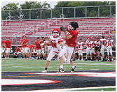 Two people in sports uniforms are running on a football field with other players and equipment visible in the background. Description generated by AI