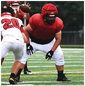 People wearing sports uniforms and cleats are positioned on a football field with artificial turf and yard lines. Description generated by AI