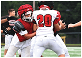 People wearing football uniforms are engaged in a play on a grassy field with one person in a red jersey and helmet attempting to tackle another in a white jersey with the number 20. Description generated by AI