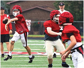 People wearing red helmets and jerseys are playing football on a field with a coach in a white shirt and black cap observing in the background. Description generated by AI
