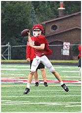 A person wearing a red football helmet and jersey is preparing to throw a football outdoors with trees and a building in the background. Description generated by AI