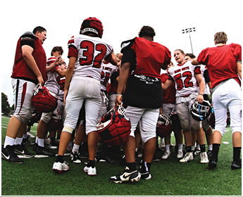 A group of people wearing football uniforms and helmets are gathered closely together on a field, appearing to be in a team huddle or discussion. Description generated by AI