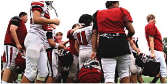 People wearing football uniforms are gathered closely together, some holding helmets, appearing to be in a team huddle or discussion. Description generated by AI