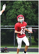 A person wearing a red sports jersey, white pants, and a black helmet is reaching out with one arm in an outdoor sports setting with trees and a fence in the background. Description generated by AI