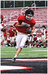 A group of people wearing football uniforms stand on the sidelines of a football field with empty bleachers in the background. Description generated by AI