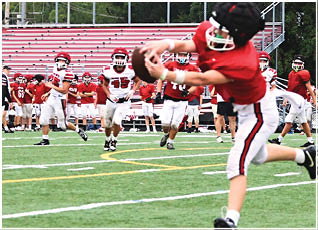 A person in red and white football gear is diving to catch a football while several other players in similar uniforms run behind on a green field with empty bleachers in the background. Description generated by AI