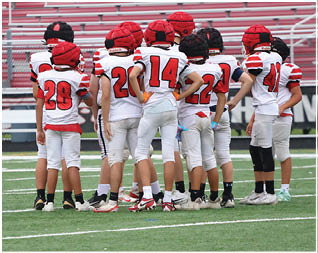 A group of children wearing football uniforms and helmets are huddled together on a football field with empty bleachers in the background. Description generated by AI