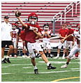 A person wearing a red helmet and jersey is throwing a football on a field with other people in sports gear in the background. Description generated by AI