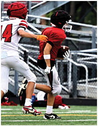 A person wearing a red sports jersey and black helmet is holding a football and running on a field with metal bleachers in the background. Description generated by AI