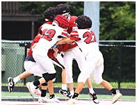 Four people wearing football helmets and uniforms are engaged in a play on a field near a chain-link fence. Description generated by AI