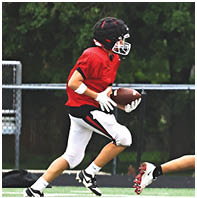 A person wearing a red sports jersey, white gloves, and a black helmet is holding a football on a field with a chain-link fence and trees in the background. Description generated by AI