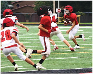 Children wearing football helmets and uniforms are playing on a grassy field with one child holding a football and preparing to run. Description generated by AI