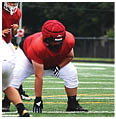A person wearing a red football helmet and jersey with white pants is crouched on a football field. Description generated by AI