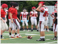 Several people in football uniforms stand on a field with a football lying on the ground in front of them. Description generated by AI