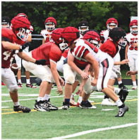 Children wearing red and white football uniforms and helmets are lined up on a grassy field preparing for a play during a game. Description generated by AI
