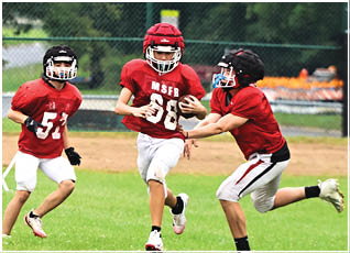 Three children wearing red football uniforms and helmets are playing on a grassy field with one child running while holding the football and another attempting to tackle. Description generated by AI