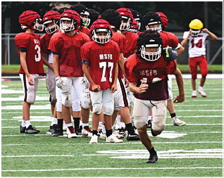 A group of children wearing red and black football helmets and jerseys are standing and preparing on a football field. Description generated by AI