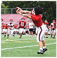 A person in a red sports uniform and helmet is reaching out to catch a football while other people in similar uniforms and a coach stand in the background near bleachers. Description generated by AI
