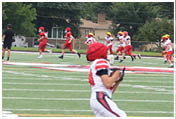 A person in a white and red sports uniform is running on a football field near the end zone while a group of people in red uniforms and coaches stand near the goalpost in the background. Description generated by AI