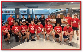 A group of people wearing red and black sports shirts pose together indoors in a cafeteria-like setting with flags hanging in the background. Description generated by AI