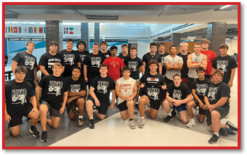 A group of adults wearing matching black sports shirts with “Hawks" printed on them are posing together indoors in a well-lit room with flags hanging in the background. Description generated by AI