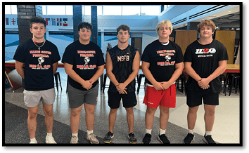 Five adults stand side by side indoors wearing casual shorts and t-shirts with sports team logos and text, posing for a group photo. Description generated by AI