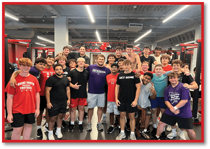 A group of adults wearing athletic clothing pose together smiling inside a gym with weightlifting equipment in the background. Description generated by AI