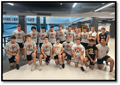 A group of adults wearing athletic clothing pose together smiling inside a gym with weightlifting equipment in the background. Description generated by AI
