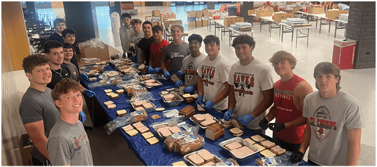 A group of people wearing casual clothing and gloves stand around a long table covered with a blue plastic tablecloth preparing sandwiches with bread and various ingredients. Description generated by AI
