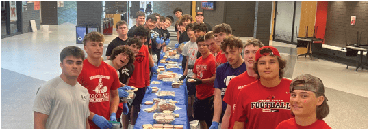 A group of people wearing casual clothing and sports-themed shirts stand on both sides of a long table covered with food trays in a large indoor space. Description generated by AI