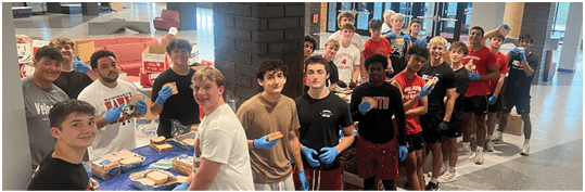 A group of people wearing casual clothing and gloves are standing in a line indoors, preparing sandwiches on a table covered with a blue cloth. Description generated by AI