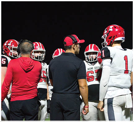 A group of people wearing football uniforms and helmets are gathered on a field at night, with two individuals in coaching attire standing among them. Description generated by AI