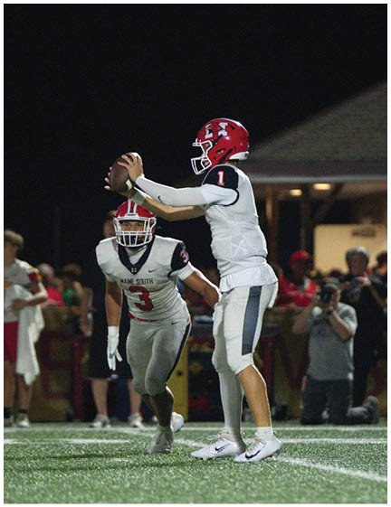 A person in a white and red football uniform is holding a football ready to pass while another person in a similar uniform runs toward them on a lit football field at night with spectators in the background. Description generated by AI