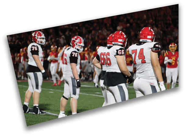 Several people wearing football uniforms stand on a field with one person in the background holding a football. Description generated by AI