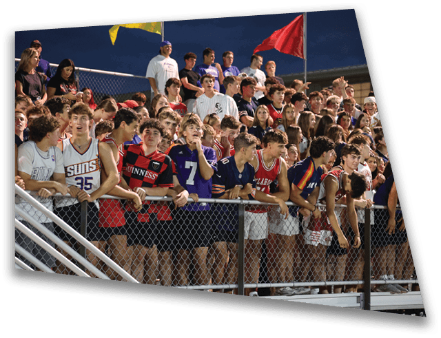 A large group of people wearing sports jerseys and casual clothing are gathered closely together behind a chain-link fence at an outdoor event during the evening. Description generated by AI