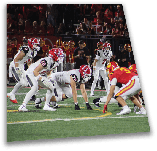 Adults in football uniforms are lined up on a field facing each other in a game setting with spectators in the background. Description generated by AI