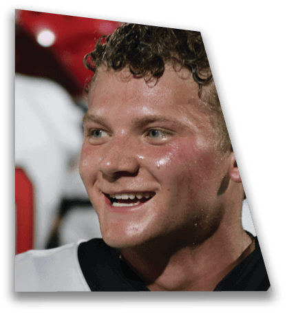 A person with curly hair is smiling and wearing a sports jersey with a blurred background showing another person in a similar jersey. Description generated by AI