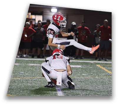 Two people in football uniforms are on a field with one holding the ball on the ground while the other kicks it. Description generated by AI