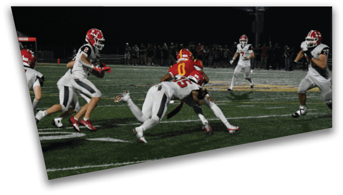 Several people in football uniforms are actively engaged in a play on a field under stadium lights, with one person tackling another wearing a red jersey. Description generated by AI