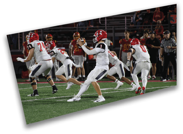 A group of people wearing football uniforms are actively playing on a field with spectators and other players visible in the background. Description generated by AI